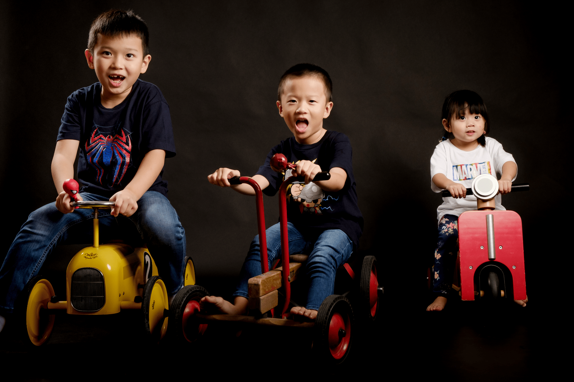 3 children pose on toy scooters against a black studio background